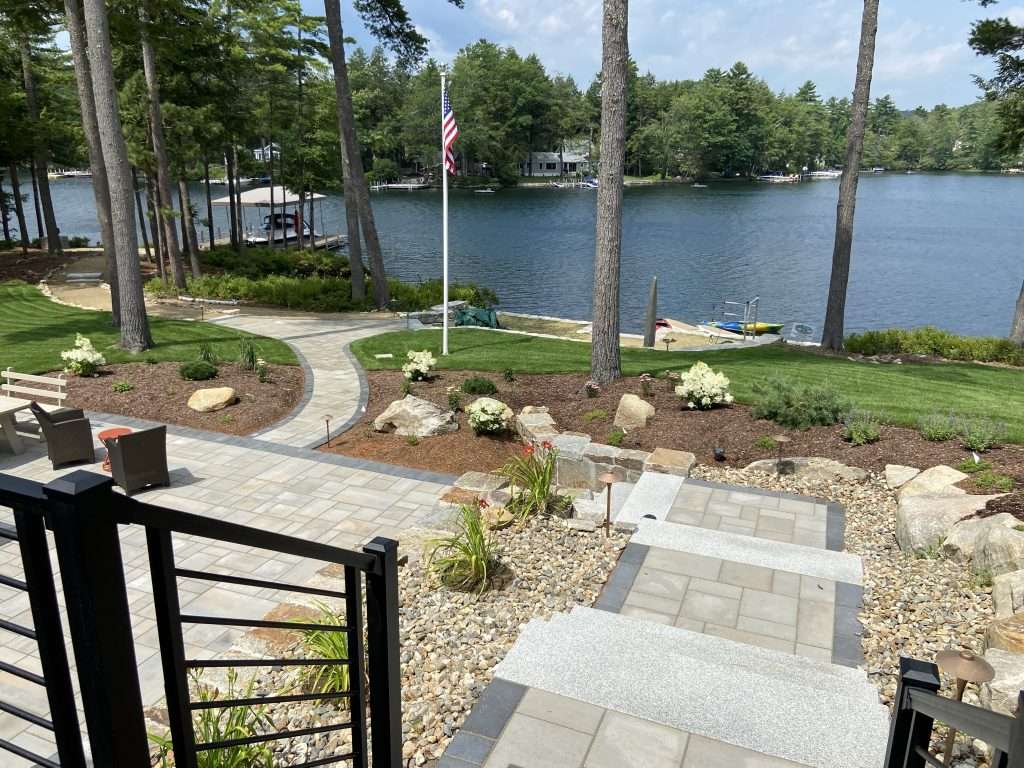 Custom granite steps and terraced hardscape installation featuring natural stone retaining walls, paver walkways, decorative boulder accents, and professionally designed landscape plantings for a New Hampshire lakeside property