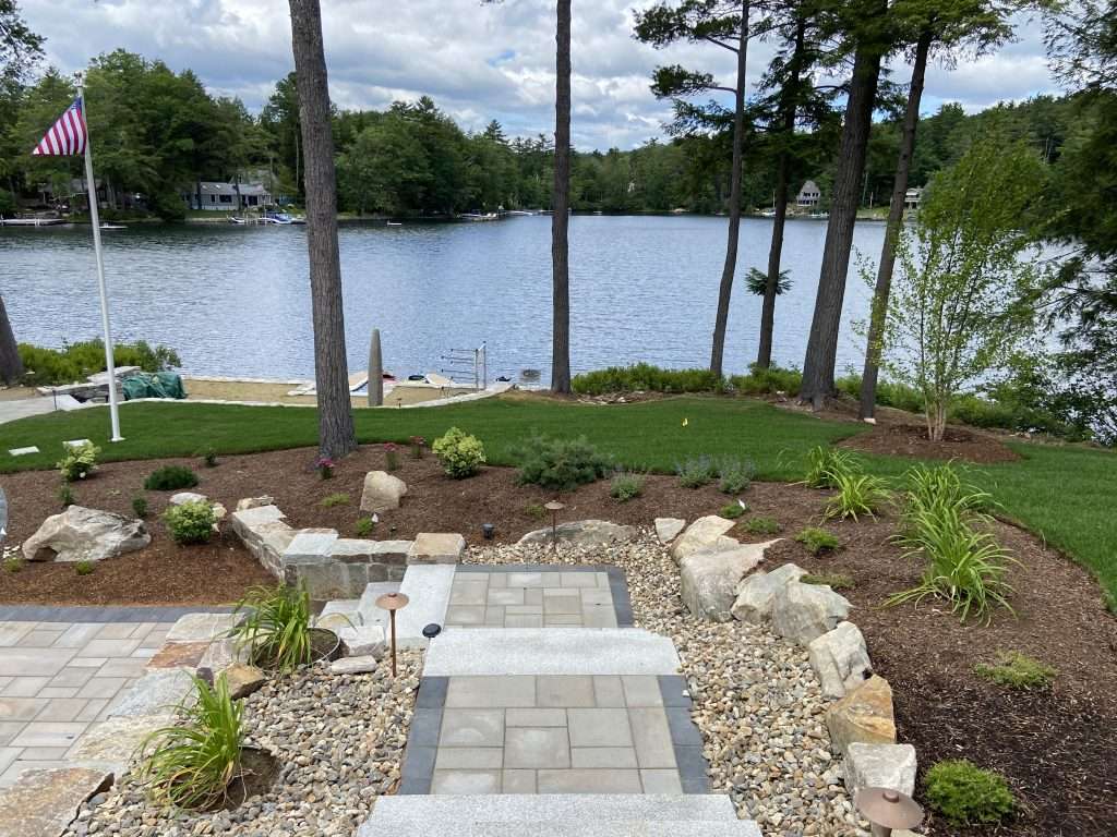 Luxury Lakes Region hardscape and landscape project featuring custom granite steps, natural stone retaining walls, paver driveway and walkways, boulder landscaping, drainage stone accents, and professionally designed planting beds for a timeless New Hampshire outdoor living space