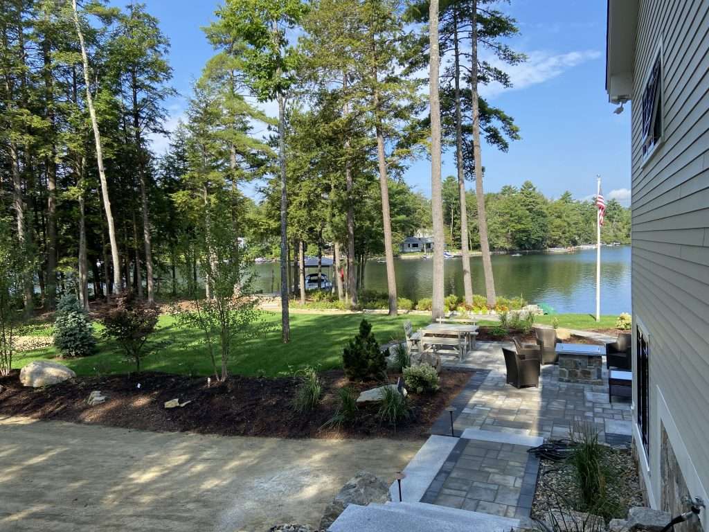 Beautiful lakefront backyard featuring custom paver patio, outdoor fire pit seating area, granite steps, natural stone accents, lush lawn installation, and professionally designed landscaping overlooking a New Hampshire lake