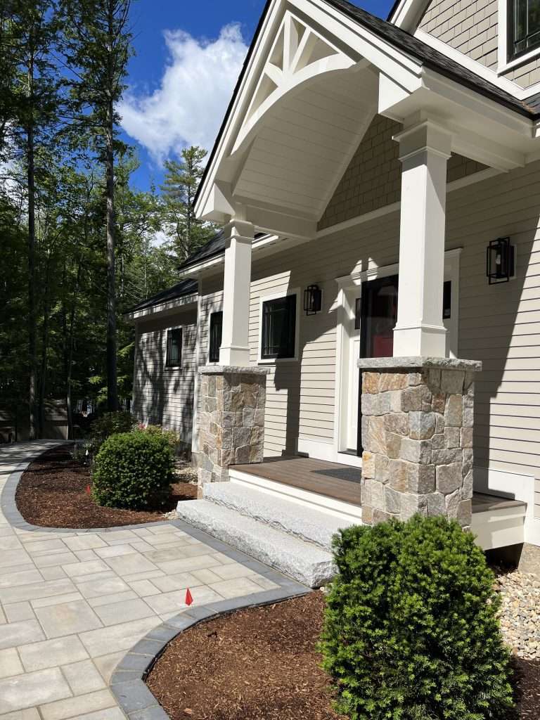 Elegant front entry hardscape with custom paver walkway, granite front steps, natural stone column veneers, curved landscape edging, and professionally designed foundation plantings for a luxury New Hampshire home