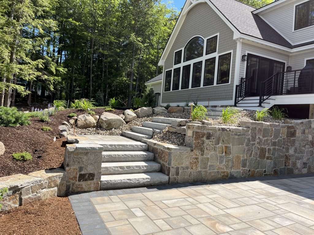 Luxury hardscape installation with custom paver patio, granite stairway, natural stone retaining wall, decorative landscape boulders, and modern outdoor living design for a New Hampshire home