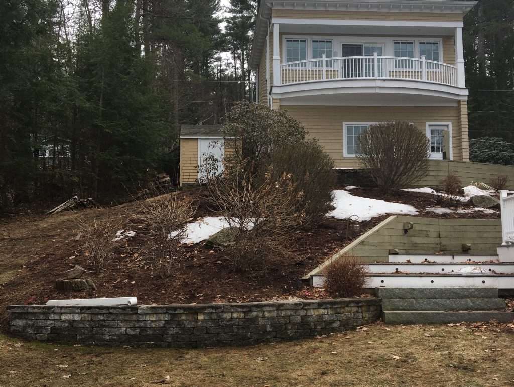 Before photo of a sloped backyard area prior to terraced patio, retaining wall, and outdoor fireplace installation by Landscapes by Tom in New Hampshire