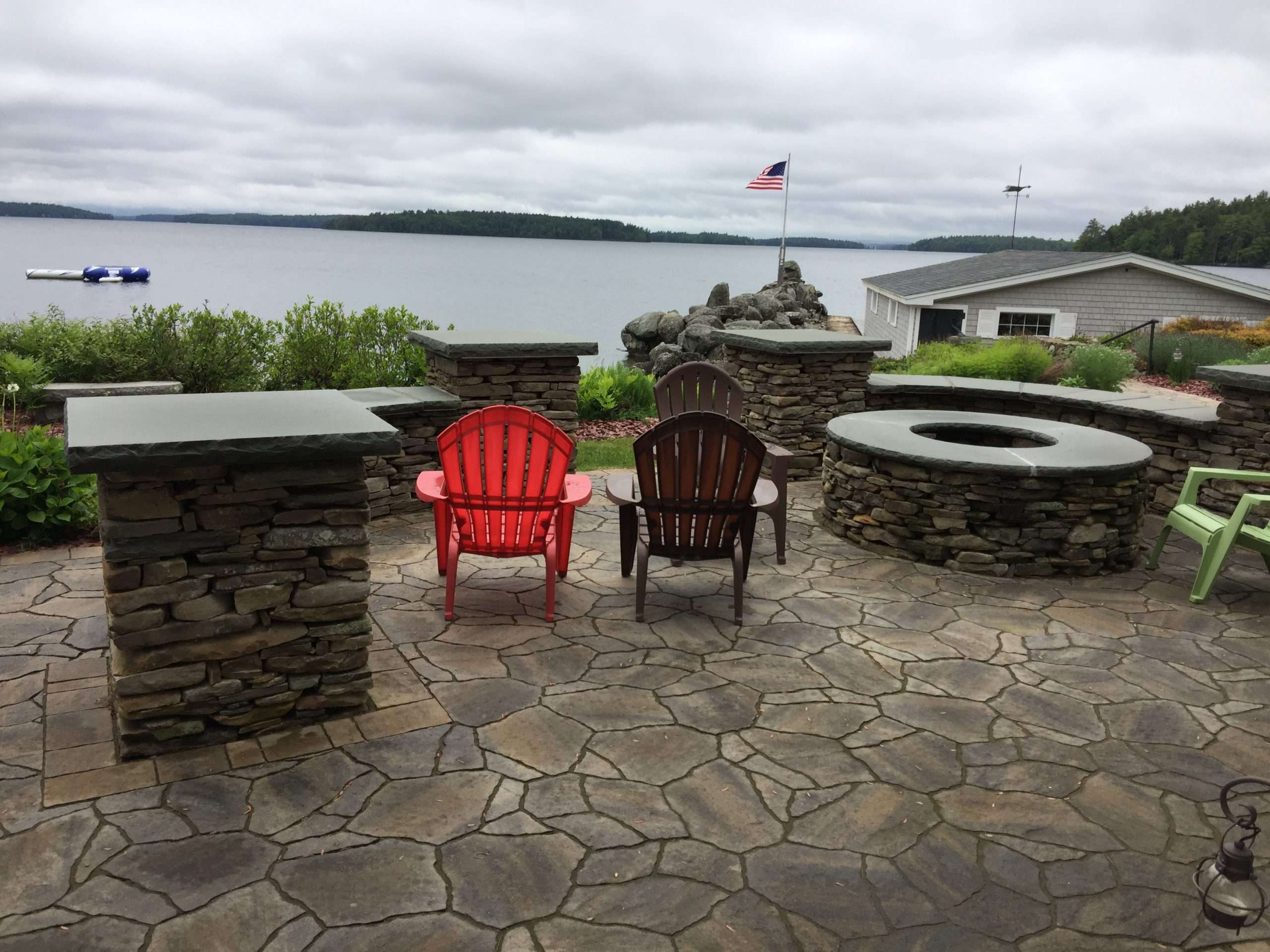 Natural stone lakeside patio with custom masonry fire pit, stacked stone seating walls, granite caps, Adirondack chairs, and panoramic waterfront views designed for outdoor entertaining and relaxation