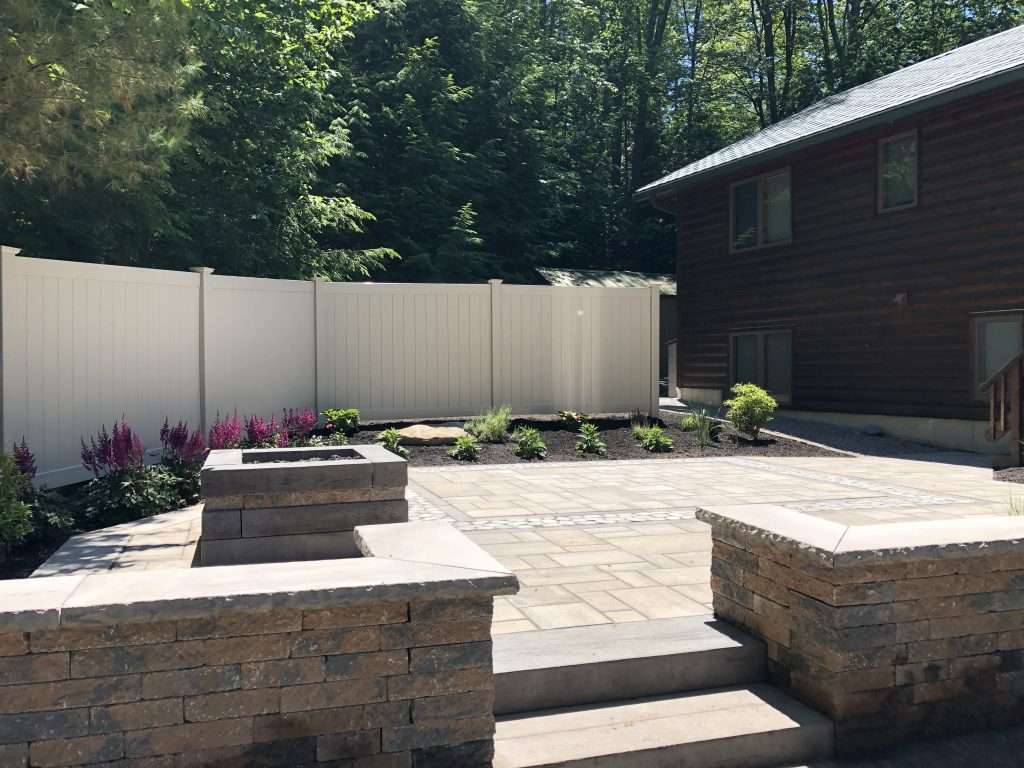 Elevated backyard patio with custom paver hardscape, built-in fire pit, stone seating walls, decorative granite steps, and landscaped planting beds in a wooded outdoor living setting