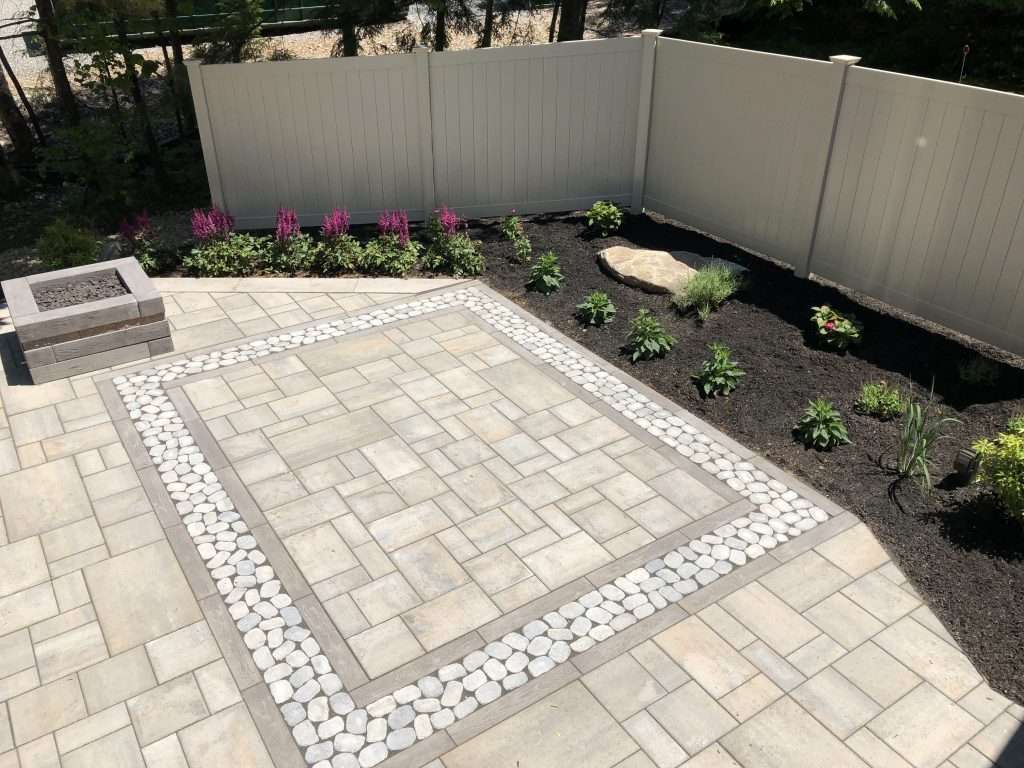 Custom paver patio with decorative river rock inlay, modern fire pit, geometric hardscape design, and landscaped garden beds for a stylish backyard outdoor living space 2