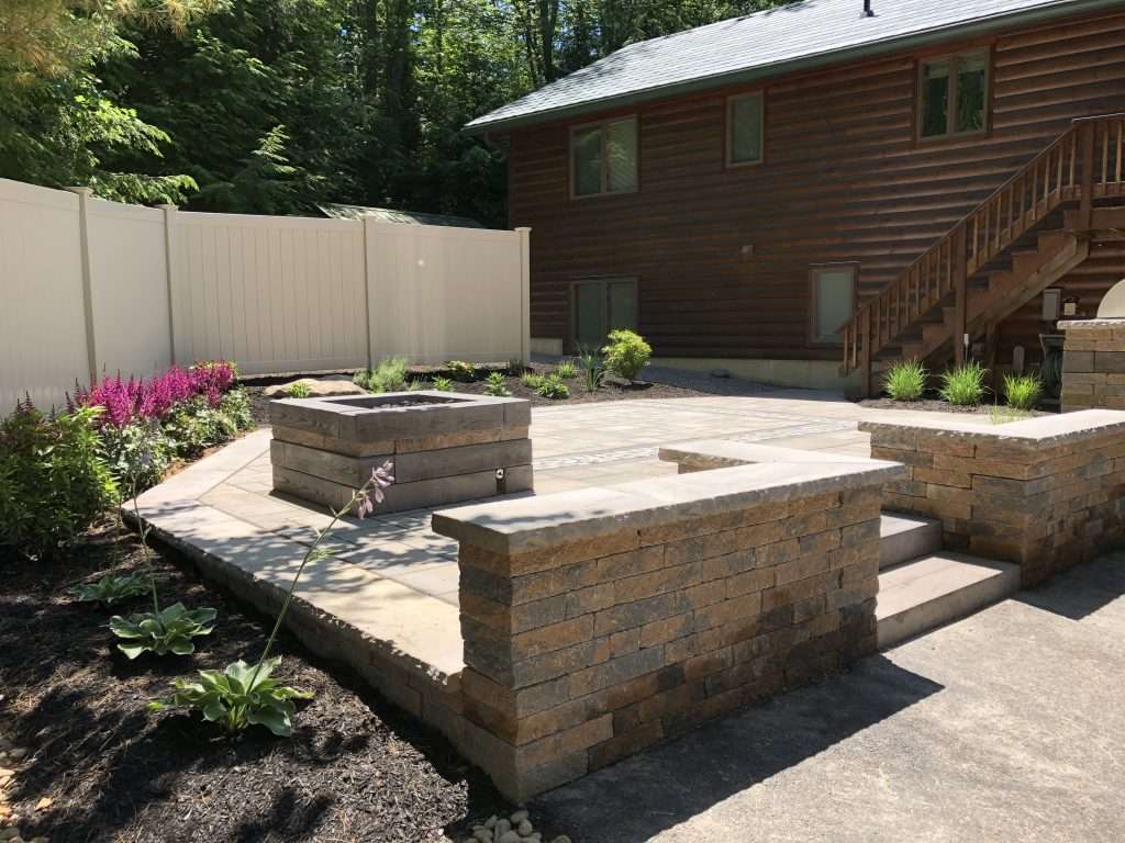 Custom hardscape patio with paver installation, built-in fire pit, retaining walls with stone caps, granite steps, and landscaped backyard outdoor living area surrounded by natural woodland