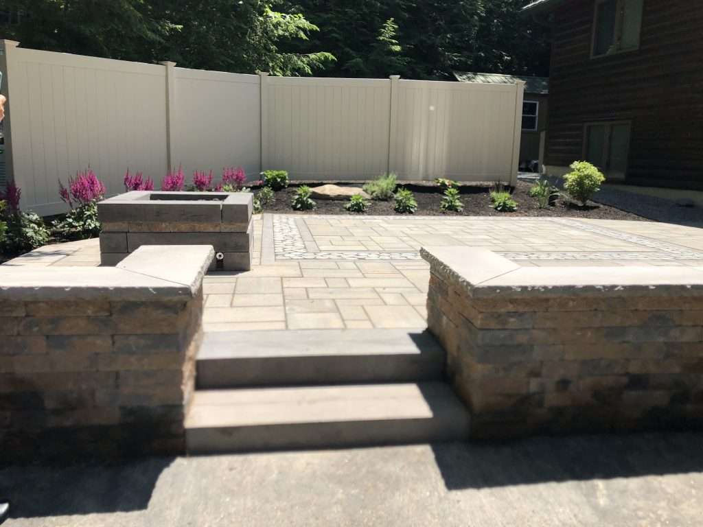 Modern backyard patio with custom paver installation, built-in fire pit, natural stone seating walls, granite steps, decorative stone accents, and professionally landscaped outdoor living space
