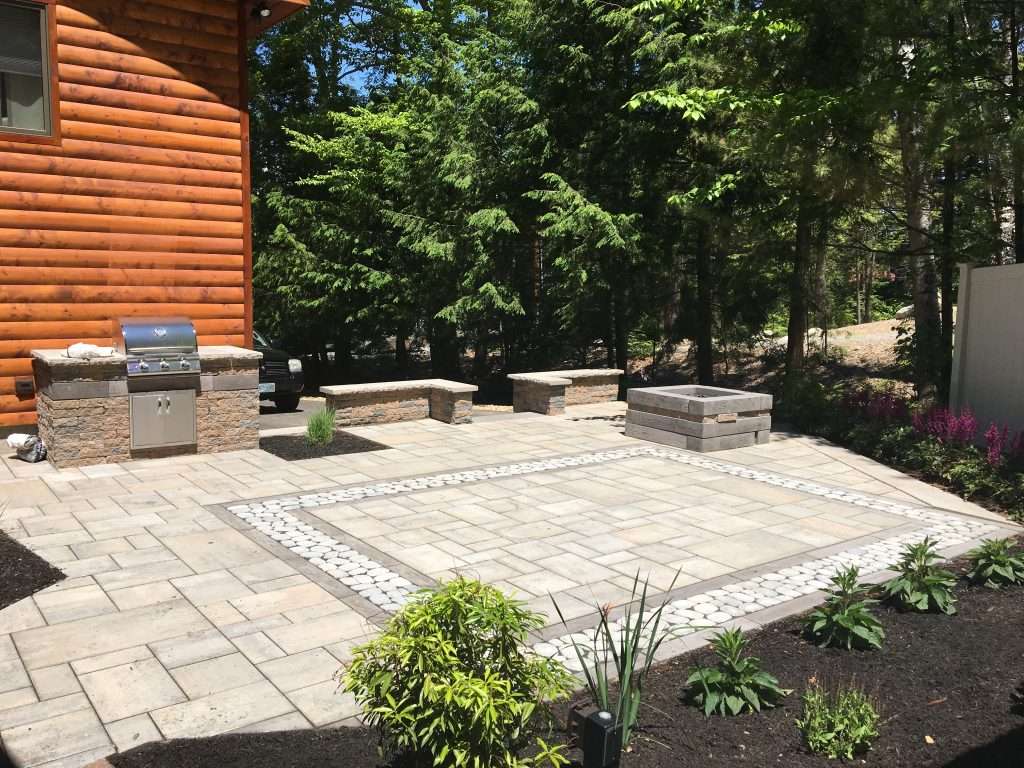 Custom backyard patio with modern paver design, built-in outdoor kitchen and grill, gas fire pit, seating walls, decorative stone inlay border, and landscaped outdoor living area in New Hampshire