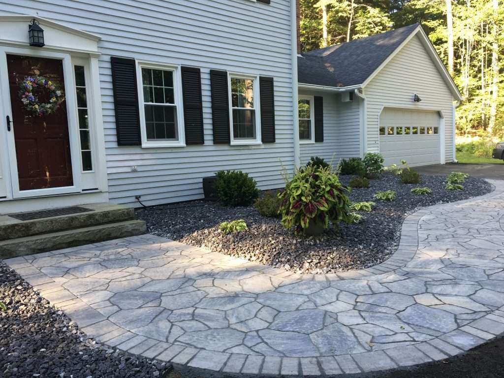 Custom natural stone walkway and front entry landscaping featuring curved hardscape design, decorative river rock beds, granite front steps, foundation plantings, and professionally crafted curb appeal enhancements for a New Hampshire home