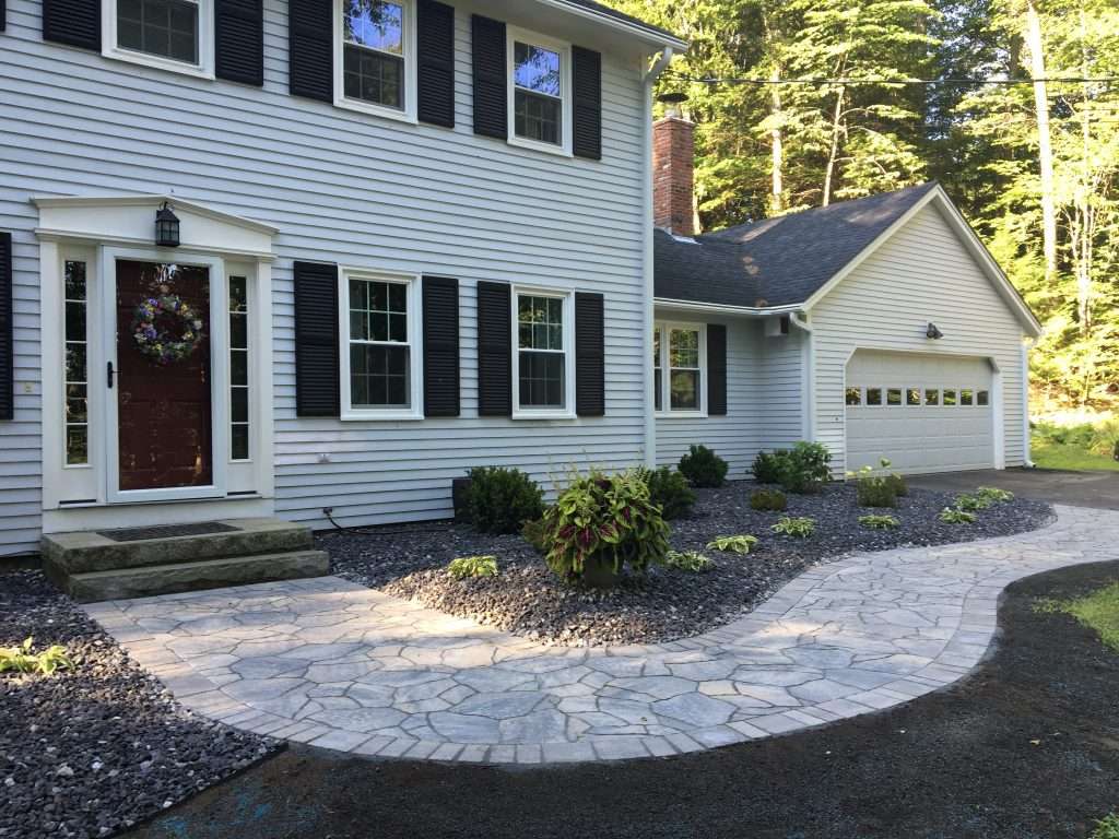 Curved natural stone walkway with decorative border leading to a residential front entrance, complemented by low-maintenance foundation landscaping, river rock beds, and clean landscape edging