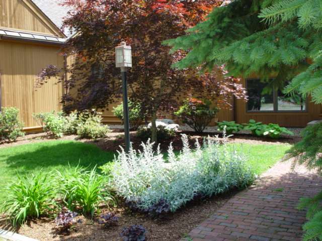Interesting front entry bed plantings featuring layered shrubs, flowering perennials, ornamental grasses, fresh mulch, and professionally designed foundation landscaping for enhanced curb appeal in New Hampshire